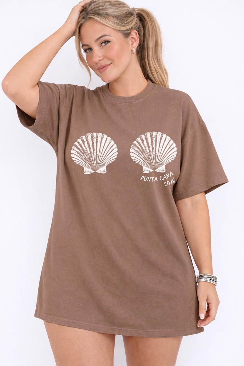 May include: A brown t-shirt featuring two white seashell graphics and the text "PUNTA CANA 2026". The shirt has short sleeves and a relaxed fit, suitable for casual wear.