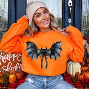 May include: An orange sweatshirt featuring a black bat wing design. The bat wings are detailed with a bow in the centre. The sweatshirt is a long-sleeved crew neck. The image is of a person wearing the sweatshirt.