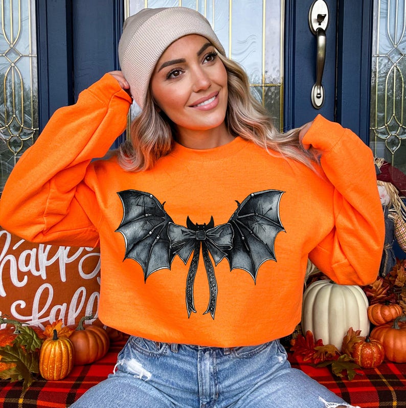 May include: An orange sweatshirt featuring a black bat wing design. The bat wings are detailed with a bow in the centre. The sweatshirt is a long-sleeved crew neck. The image is of a person wearing the sweatshirt.