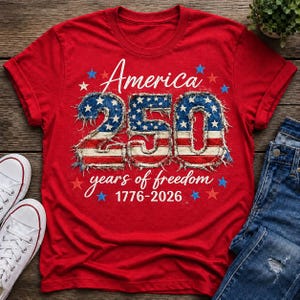 May include: A red t-shirt with the text "America 250 years of freedom 1776-2026" in white script. The numbers "250" are designed with a distressed American flag pattern, surrounded by blue stars. A pair of white sneakers and blue jeans are visible.