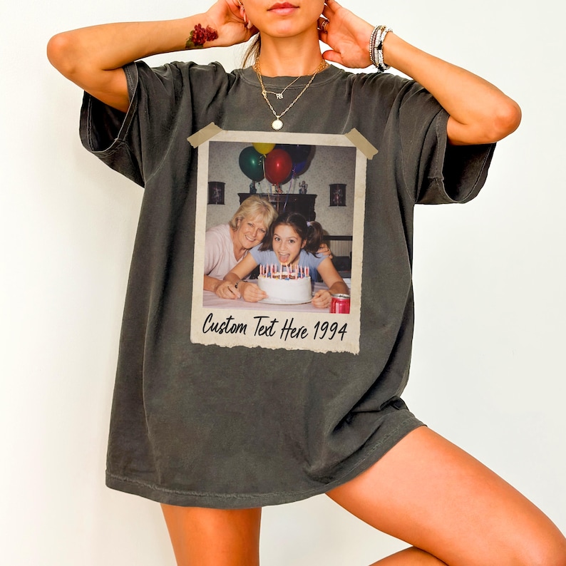 Puede incluir: Camiseta gris oscuro con estampado fotogr&aacute;fico de estilo vintage. La foto muestra a dos personas sonriendo frente a una tarta de cumplea&ntilde;os con velas. El texto "Custom Text Here 1994" est&aacute; debajo. La camiseta tiene mangas cortas.