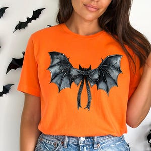 May include: An orange t-shirt with a black and grey bat wing design and a bow. The graphic is in the centre of the shirt. The background features black bat decorations. The t-shirt is a solid orange colour.