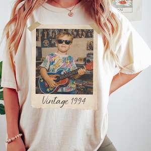 Puede incluir: Camiseta color crema con estampado de foto vintage. La foto muestra a una persona con gafas de sol tocando un ukelele. La foto lleva la leyenda "Vintage 1994".