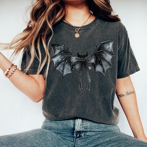 May include: Dark grey t-shirt with a bat wing and bow design. The wings are detailed with shading and texture, and the bow has long, flowing ribbons. The shirt has a casual style and a slightly faded appearance.
