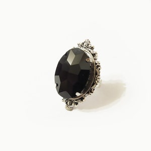 Può includere: Un anello d'argento con una grande pietra preziosa nera sfaccettata incastonata in una cornice decorata e ornata.