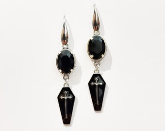 May include: A pair of silver dangle earrings with black oval-shaped stones and a coffin charm with a cross on each earring.