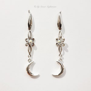 Può includere: Orecchini pendenti in argento con un ciondolo a forma di luna crescente e una piccola pietra iridescente in un design floreale.