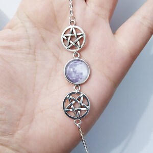 May include: A silver chain necklace with three charms: a pentagram, a moon, and another pentagram. The moon charm has a purple background with a white moon image.