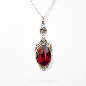 May include: A silver-toned pendant necklace with an oval, deep red gemstone. The gemstone is set in an ornate, filigree-style frame. The necklace hangs from a delicate chain. The text "oMySweetNightmareOo" is visible.