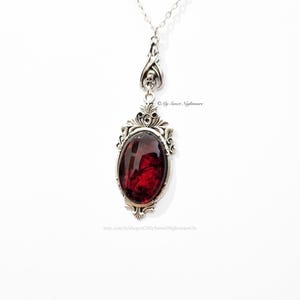 May include: A silver-toned pendant necklace featuring an oval, deep red gemstone set in an ornate frame. The pendant hangs from a delicate chain. The design includes decorative flourishes and a skull detail. The Etsy shop name "My Sweet Nightmare" is visible.