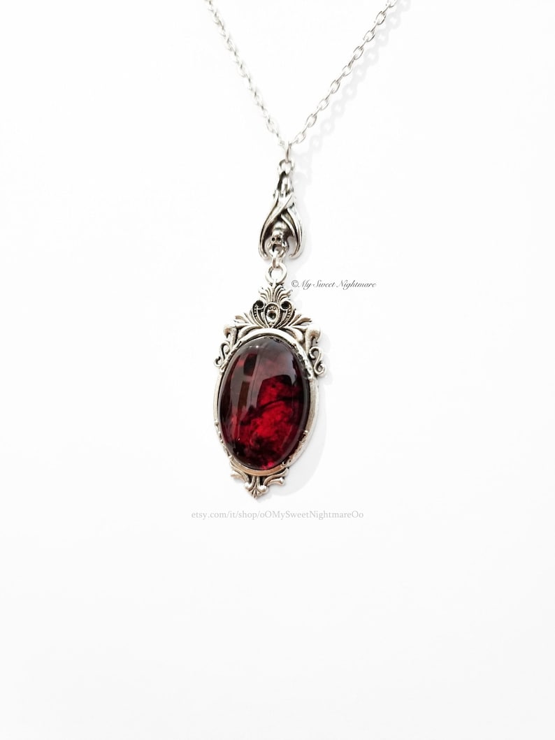 May include: A silver-toned pendant necklace featuring an oval, deep red gemstone set in an ornate frame. The pendant hangs from a delicate chain. The design includes decorative flourishes and a skull detail. The Etsy shop name "My Sweet Nightmare" is visible.