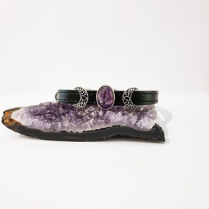 May include: A black leather bracelet with silver crescent moon charms and a purple amethyst stone in the center.