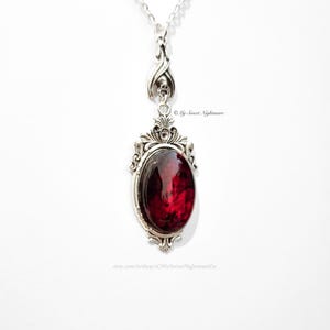 May include: Silver-toned pendant necklace with an oval, deep red gemstone. The pendant has an ornate, gothic-style frame and a decorative bail with a skull detail. The necklace is on a white background.
