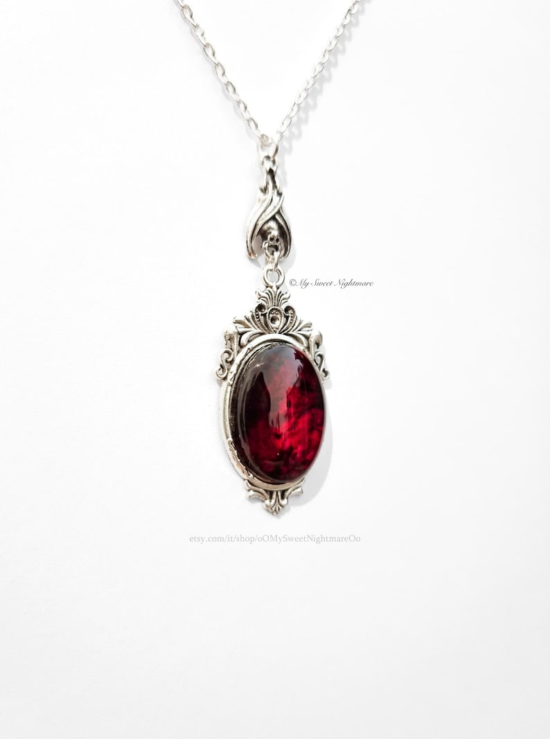 May include: Silver-toned pendant necklace with an oval, deep red gemstone. The pendant has an ornate, gothic-style frame and a decorative bail with a skull detail. The necklace is on a white background.