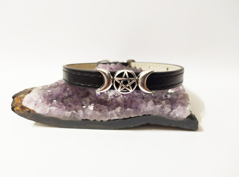 May include: A black leather choker necklace with a silver triple moon and pentagram charm. The necklace is resting on a purple amethyst crystal.