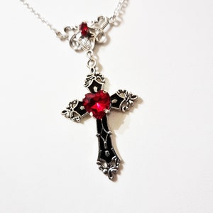 Puede incluir: Collar plateado con un colgante de cruz negra con una gema roja en forma de coraz&oacute;n. La cruz tiene detalles ornamentados y cuelga de una delicada cadena. Una gema roja m&aacute;s peque&ntilde;a adorna la parte superior del collar.