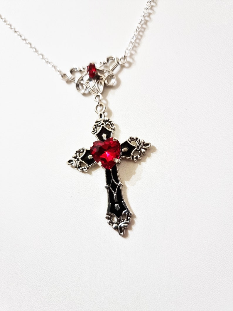 Puede incluir: Collar plateado con un colgante de cruz negra con una gema roja en forma de coraz&oacute;n. La cruz tiene detalles ornamentados y cuelga de una delicada cadena. Una gema roja m&aacute;s peque&ntilde;a adorna la parte superior del collar.