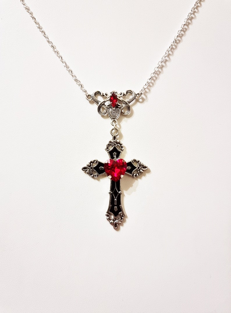 Puede incluir: Collar plateado con un colgante de cruz g&oacute;tica con una gema roja en forma de coraz&oacute;n en el centro. La cruz tiene detalles negros y ornamentados. Una gema roja m&aacute;s peque&ntilde;a est&aacute; engastada encima de la cruz. Fondo blanco.