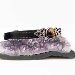 K&ouml;nnte beinhalten: Schwarzes Lederhalsband mit silberfarbenen keltischen Knoten und einem zentralen Tigeraugenstein. Das Halsband wird auf einer lila-schwarzen Geode pr&auml;sentiert, was eine mystische &Auml;sthetik zeigt.