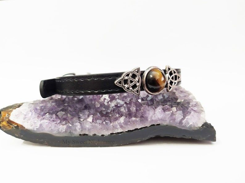 K&ouml;nnte beinhalten: Schwarzes Lederhalsband mit silberfarbenen keltischen Knoten und einem zentralen Tigeraugenstein. Das Halsband wird auf einer lila-schwarzen Geode pr&auml;sentiert, was eine mystische &Auml;sthetik zeigt.