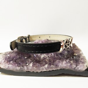 May include: A black leather choker necklace with a silver triple moon and pentacle charm. The necklace is displayed on a bed of amethyst crystals.