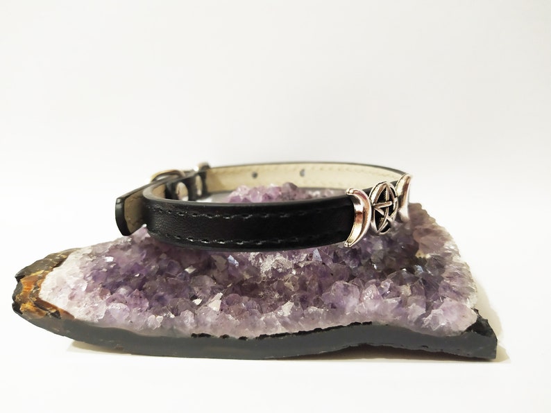 May include: A black leather choker necklace with a silver triple moon and pentacle charm. The necklace is displayed on a bed of amethyst crystals.