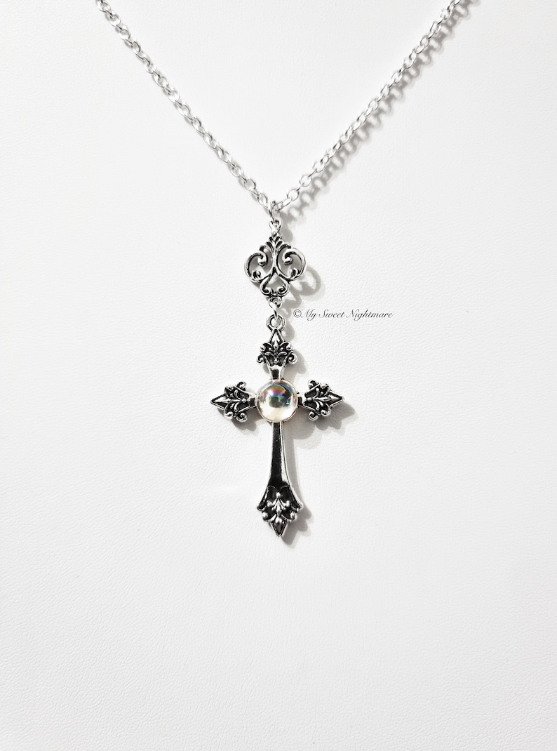 Può includere: Una collana con catena d'argento con un ciondolo a forma di croce d'argento con una perla iridescente al centro. La croce presenta intricati disegni floreali.