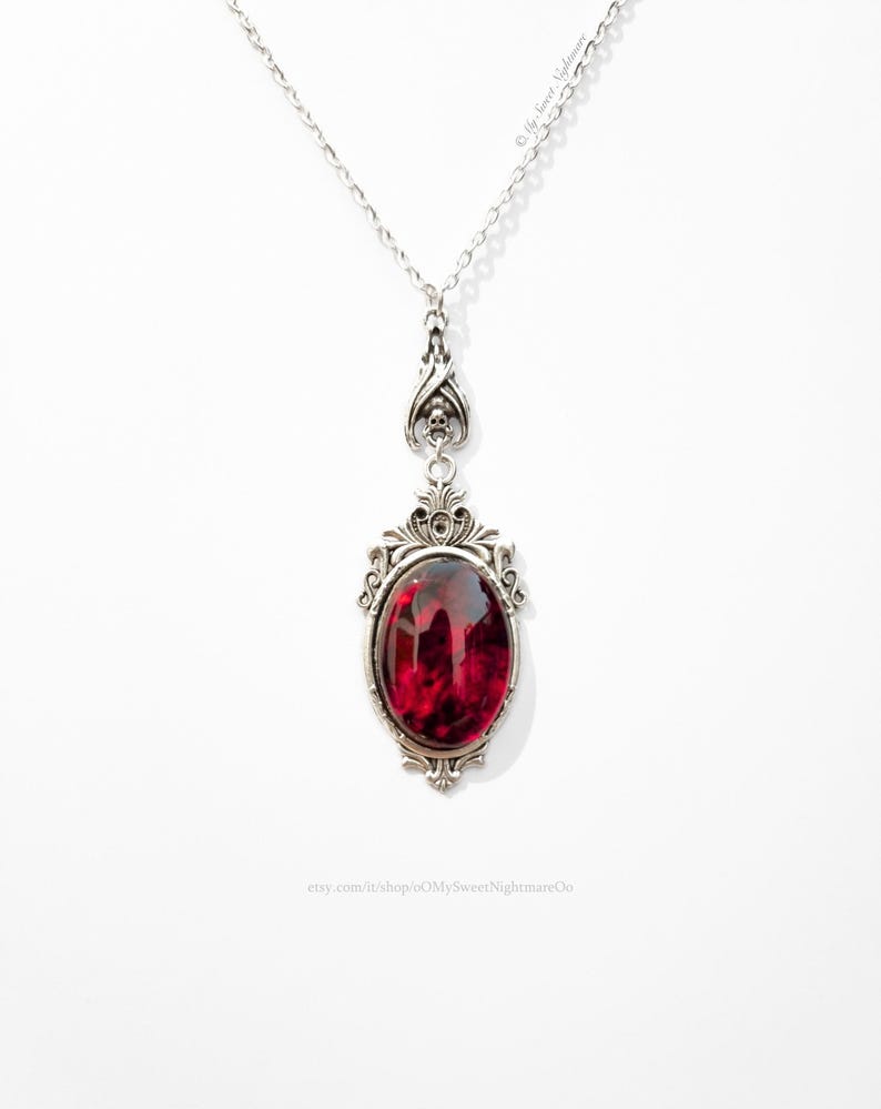 May include: A silver-toned pendant necklace with an oval, deep red gemstone. The gemstone is set in an ornate, filigree-style frame. The necklace hangs from a delicate chain. The text "oMySweetNightmareOo" is visible.