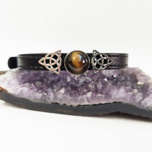 K&ouml;nnte beinhalten: Ein schwarzes Leder-Choker-Halsband mit einem zentralen Tigeraugenstein und silberfarbenen keltischen Knoten-Akzenten. Das Choker-Halsband wird auf einer lila Kristallformation pr&auml;sentiert. Das Design ist schlicht und elegant.