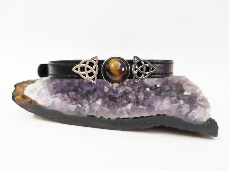 K&ouml;nnte beinhalten: Ein schwarzes Leder-Choker-Halsband mit einem zentralen Tigeraugenstein und silberfarbenen keltischen Knoten-Akzenten. Das Choker-Halsband wird auf einer lila Kristallformation pr&auml;sentiert. Das Design ist schlicht und elegant.
