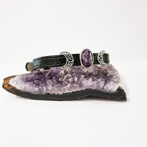 May include: A black leather bracelet with three silver crescent moon charms and a purple amethyst stone in the center.