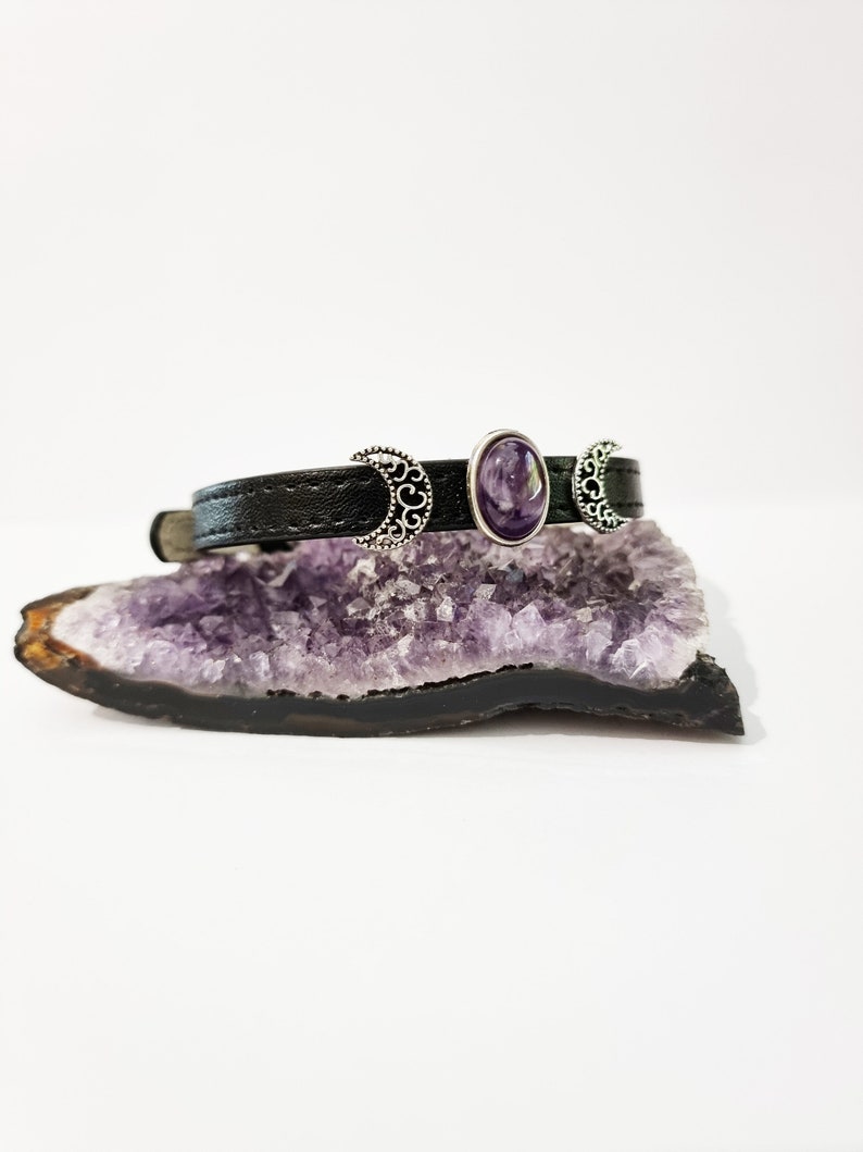 May include: A black leather bracelet with three silver crescent moon charms and a purple amethyst stone in the center.