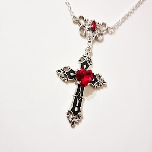 Puede incluir: Collar plateado con un colgante de cruz con una gran gema roja en forma de coraz&oacute;n en el centro. La cruz es negra con detalles plateados. Una gema roja m&aacute;s peque&ntilde;a adorna la parte superior del collar.