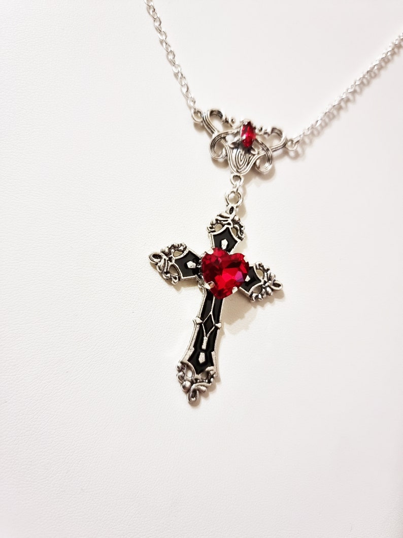 Puede incluir: Collar plateado con un colgante de cruz con una gran gema roja en forma de coraz&oacute;n en el centro. La cruz es negra con detalles plateados. Una gema roja m&aacute;s peque&ntilde;a adorna la parte superior del collar.