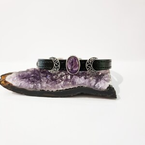 May include: A black leather bracelet with three silver crescent moon charms and a purple amethyst stone in the center.