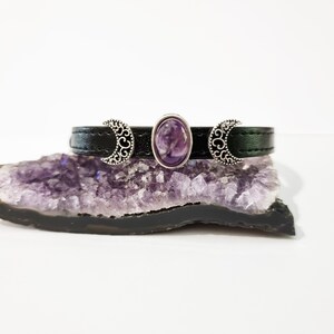 May include: A black leather bracelet with three silver crescent moon charms and a purple amethyst stone in the center.