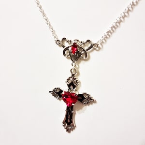 Puede incluir: Collar plateado con un colgante de cruz con una gema roja en forma de coraz&oacute;n en el centro. La cruz tiene detalles negros y ornamentados. Tambi&eacute;n hay una gema roja m&aacute;s peque&ntilde;a.