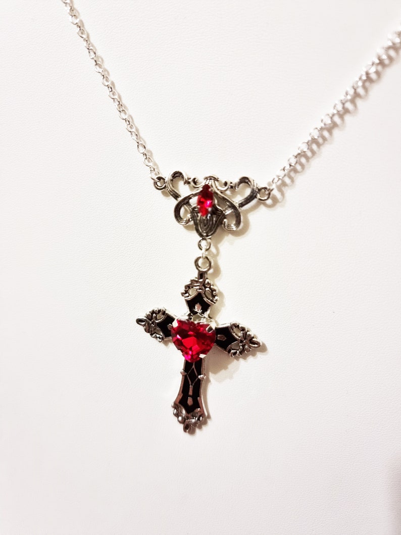 Puede incluir: Collar plateado con un colgante de cruz con una gema roja en forma de coraz&oacute;n en el centro. La cruz tiene detalles negros y ornamentados. Tambi&eacute;n hay una gema roja m&aacute;s peque&ntilde;a.