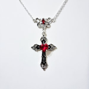 Puede incluir: Un collar plateado con un colgante de cruz. La cruz presenta una gema roja en forma de coraz&oacute;n en el centro y est&aacute; acentuada con esmalte negro. El collar tiene una cadena delicada y detalles ornamentados.