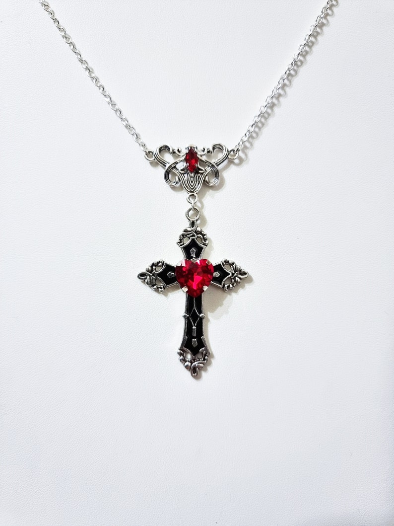 Puede incluir: Un collar plateado con un colgante de cruz. La cruz presenta una gema roja en forma de coraz&oacute;n en el centro y est&aacute; acentuada con esmalte negro. El collar tiene una cadena delicada y detalles ornamentados.