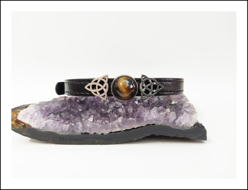 Può includere: Choker in pelle nera con dettagli a nodo celtico color argento e una pietra centrale occhio di tigre. Il choker è esposto su una geode di ametista viola, con il testo "My Sweet Nightmare" visibile.