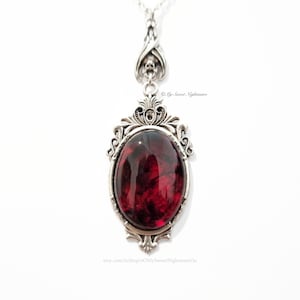 May include: Silver-toned pendant necklace with an oval, deep red stone. The stone is set in an ornate, filigree frame. The necklace has a delicate chain and a decorative bail with a skull design. The Etsy shop name is visible.