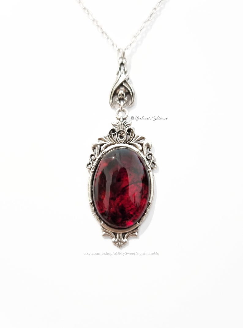 May include: Silver-toned pendant necklace with an oval, deep red stone. The stone is set in an ornate, filigree frame. The necklace has a delicate chain and a decorative bail with a skull design. The Etsy shop name is visible.