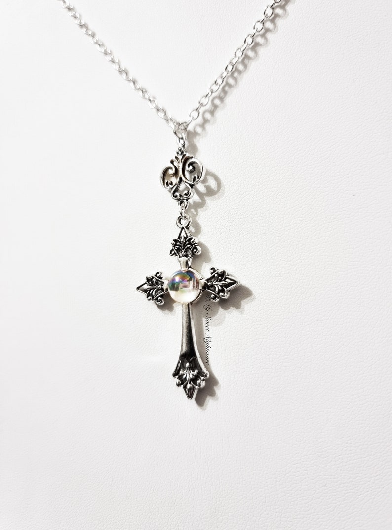 Pu&ograve; includere: Una collana con catena d'argento con un ciondolo a forma di croce d'argento con un centro in vetro iridescente.