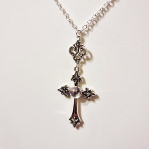 Pu&ograve; includere: Una collana a catena d'argento con un ciondolo a forma di croce d'argento. La croce ha una perla di vetro iridescente al centro.