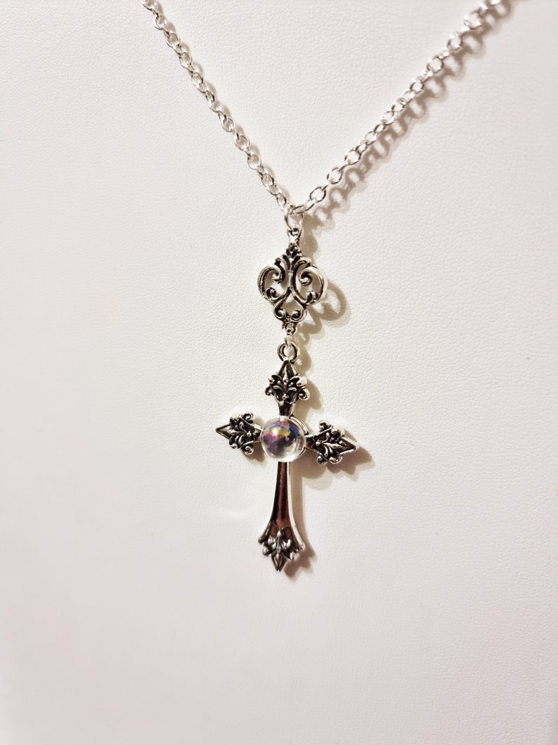 Pu&ograve; includere: Una collana a catena d'argento con un ciondolo a forma di croce d'argento. La croce ha una perla di vetro iridescente al centro.