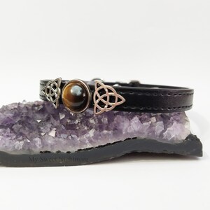 K&ouml;nnte beinhalten: Schwarzes Lederarmband mit silberfarbenen keltischen Knotenanh&auml;ngern und einem zentralen Tigeraugenstein. Das Armband wird auf einer lila Amethyst-Geode pr&auml;sentiert.