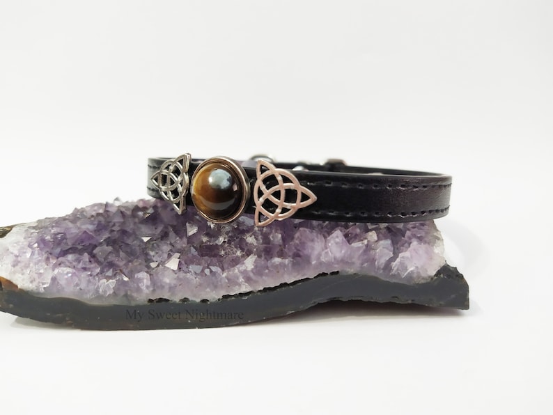 K&ouml;nnte beinhalten: Schwarzes Lederarmband mit silberfarbenen keltischen Knotenanh&auml;ngern und einem zentralen Tigeraugenstein. Das Armband wird auf einer lila Amethyst-Geode pr&auml;sentiert.
