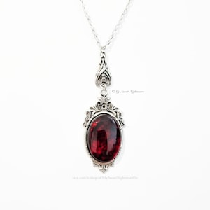 May include: Silver-toned pendant necklace featuring an oval, deep red gemstone. The pendant has an ornate, gothic-style frame with skull accents. The necklace is suspended from a delicate chain. The text "&copy; My Sweet Nightmare" is visible.