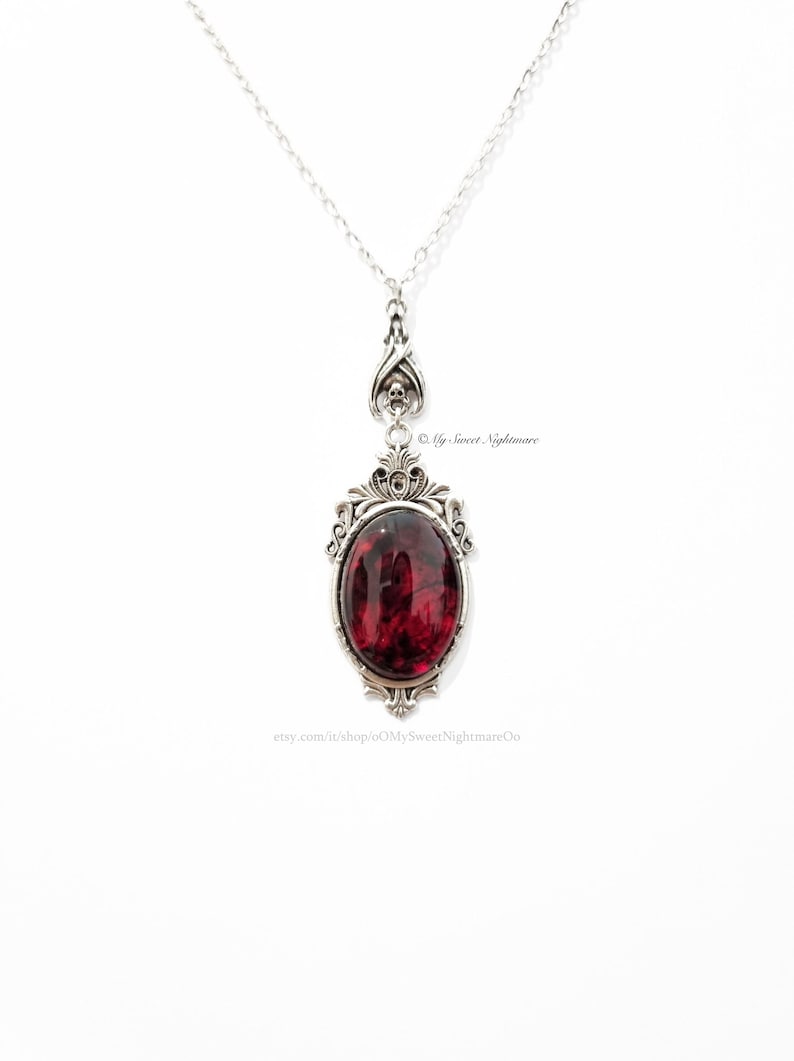 May include: Silver-toned pendant necklace with a large, oval, deep red stone. The pendant has an ornate, gothic-style frame and a delicate chain. The design includes skull accents. The Etsy shop name "oMySweetNightmareOo" is visible.